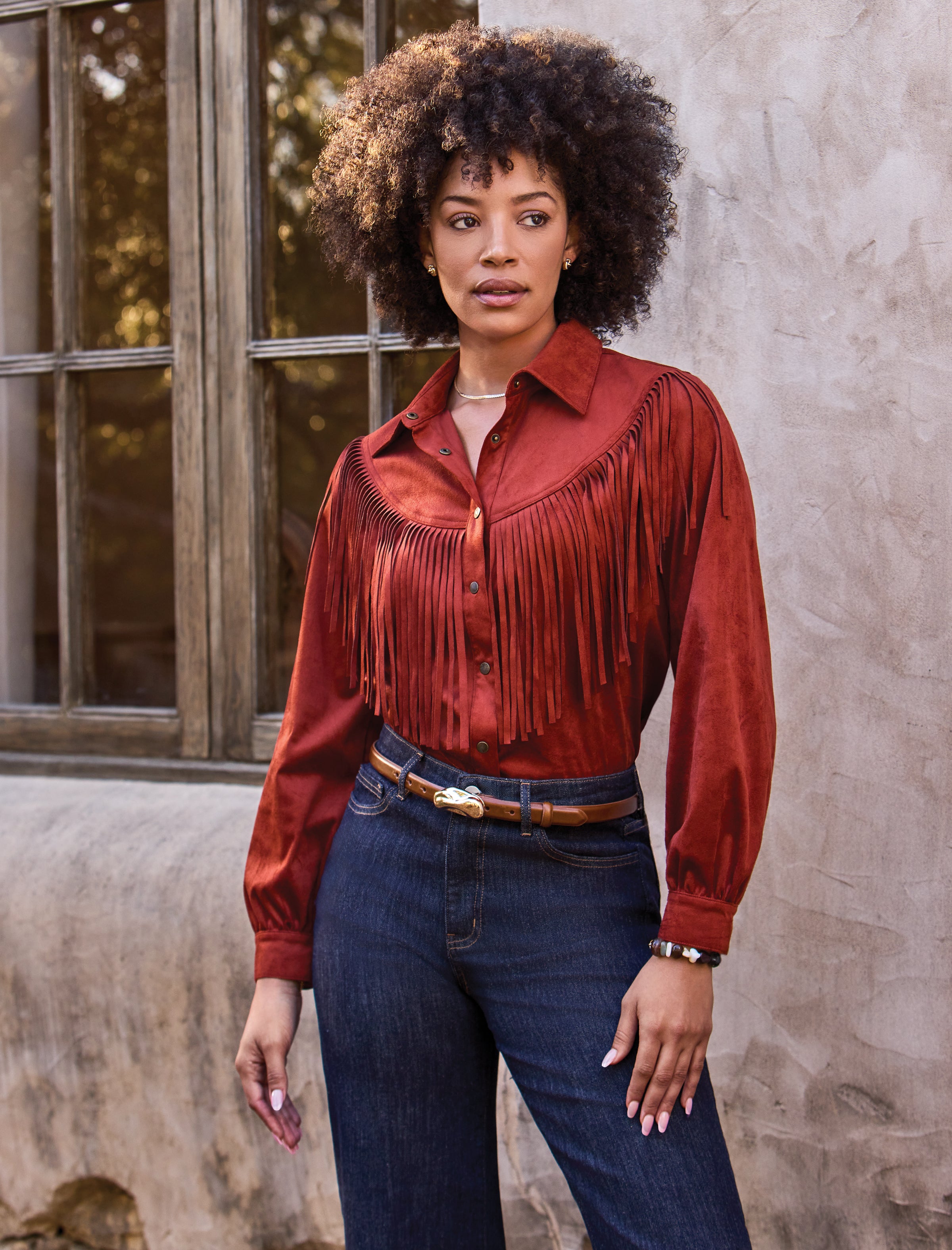 A woman with an afro hairstyle models a rust-colored long-sleeve shirt with a wide fringed yoke, paired with dark blue jeans and a brown belt, standing against a textured stucco wall and rustic window.