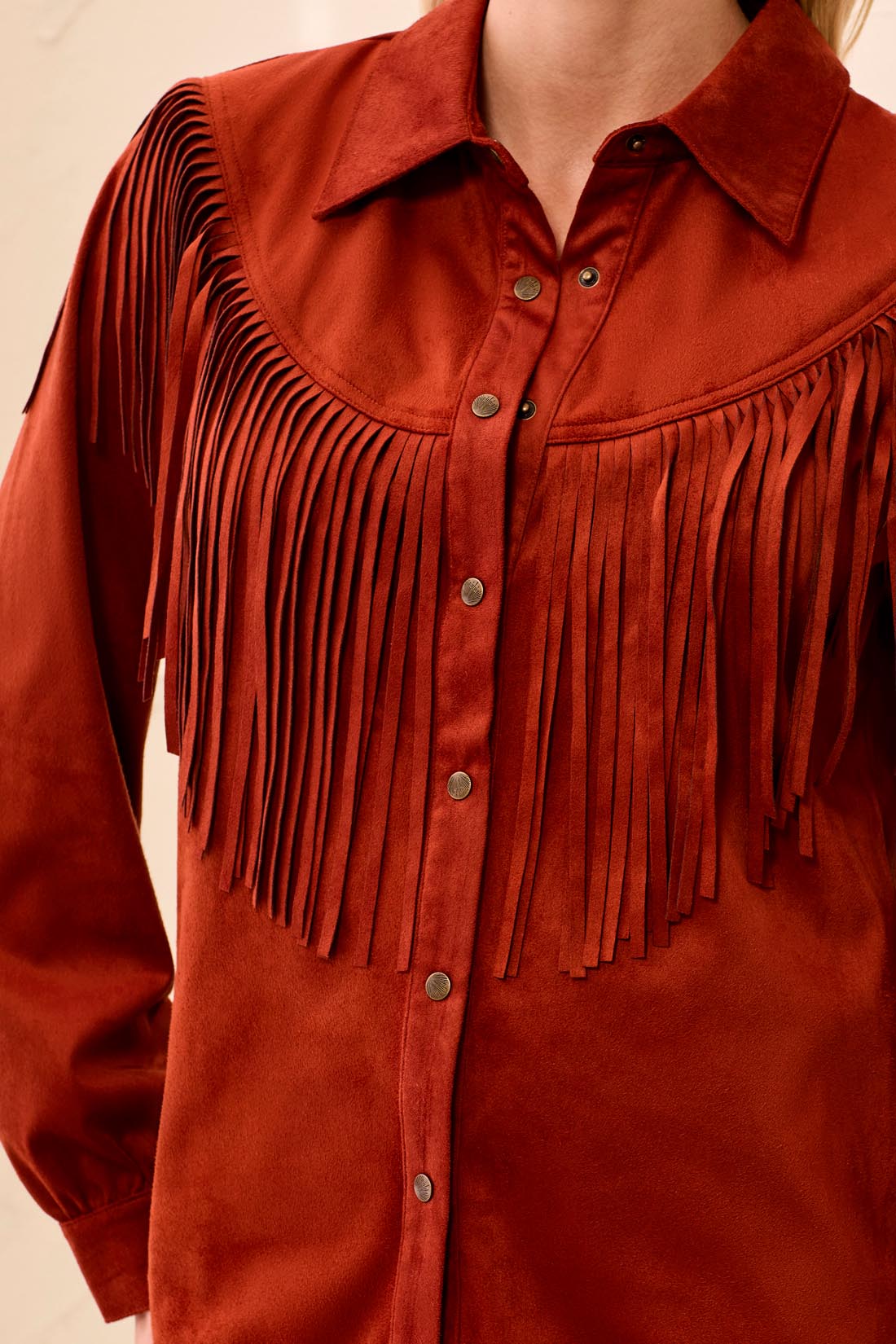 A close-up of a rust-orange suede western-style shirt jacket with a pointed collar, button front, and long fringe across the chest and shoulders.