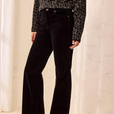 A model wearing black wide-leg corduroy pants, a black and brown leopard print long-sleeve top, and dark brown heeled ankle boots, standing in a brightly lit room with a light beige wall and wood floor.