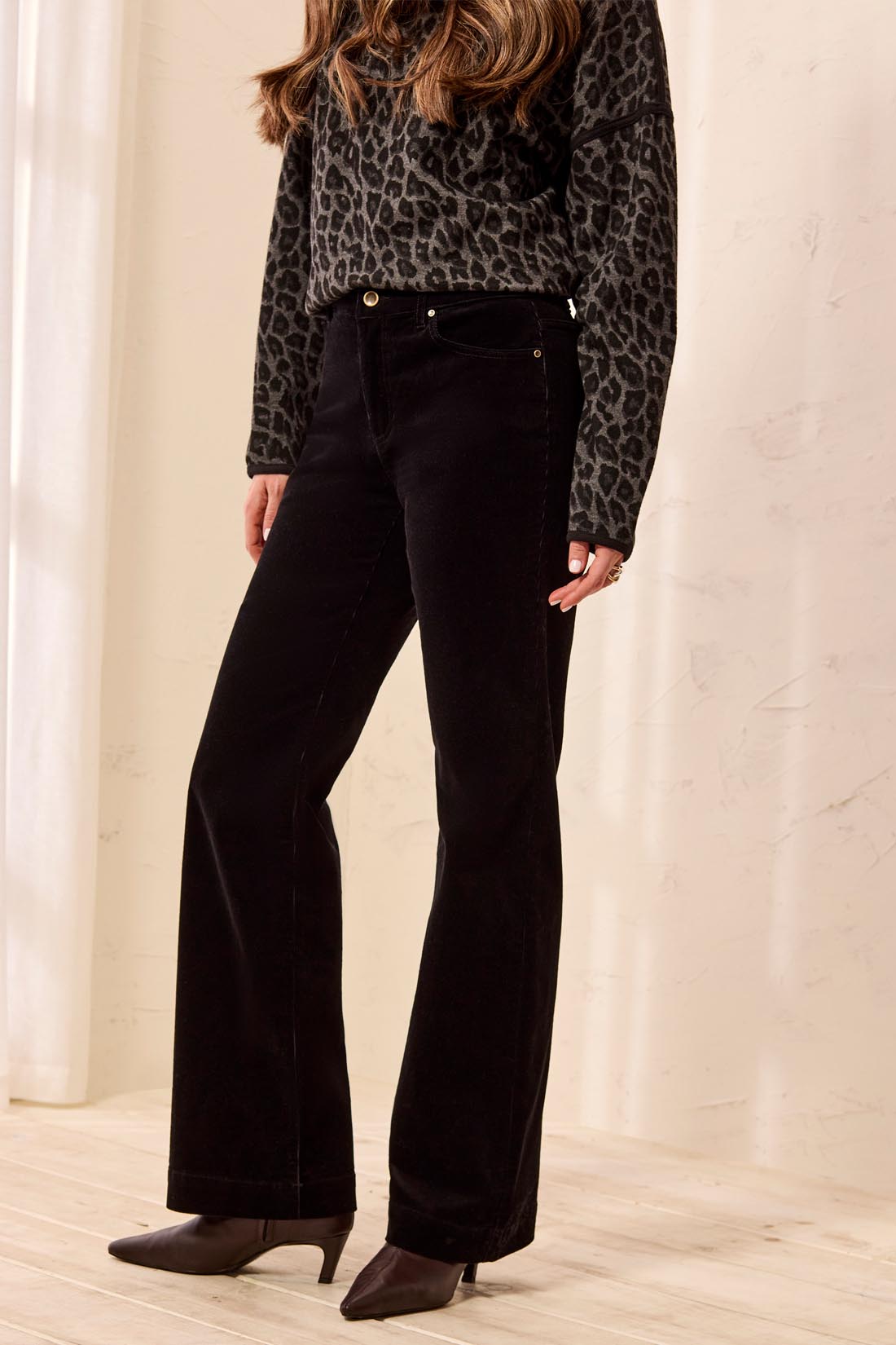 A model wearing black wide-leg corduroy pants, a black and brown leopard print long-sleeve top, and dark brown heeled ankle boots, standing in a brightly lit room with a light beige wall and wood floor.