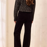 A woman shown from the back wearing a black and gray leopard-print long-sleeve crop top and black wide-leg pants, standing in a softly lit indoor setting with a light beige wall and floor.