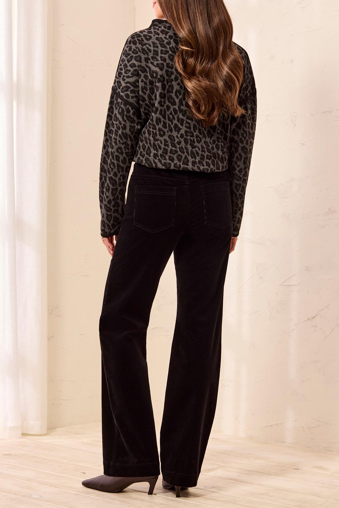 A woman shown from the back wearing a black and gray leopard-print long-sleeve crop top and black wide-leg pants, standing in a softly lit indoor setting with a light beige wall and floor.