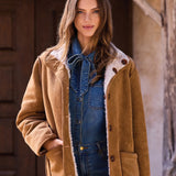 Woman wearing a brown shearling coat standing in front of a rustic building.