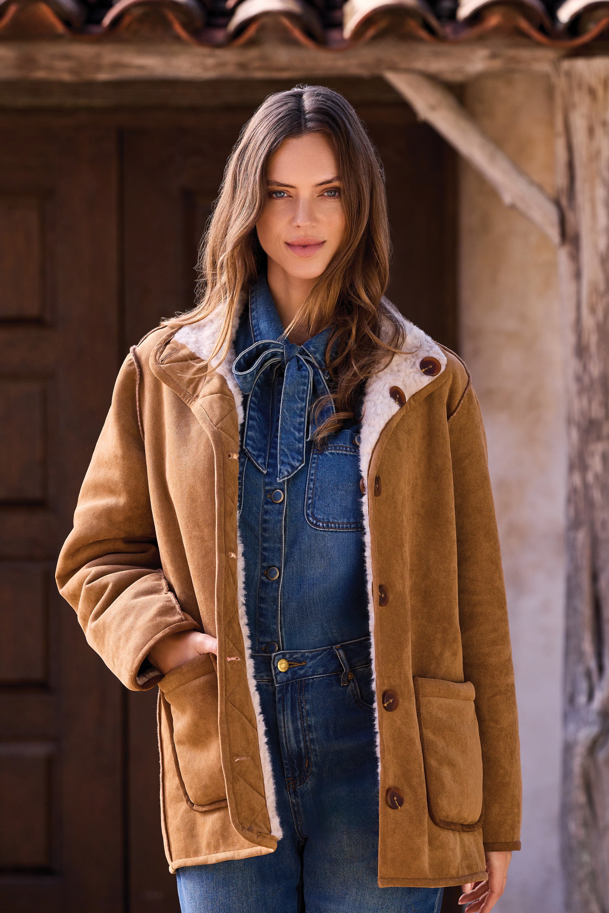 Woman wearing a brown shearling coat standing in front of a rustic building.