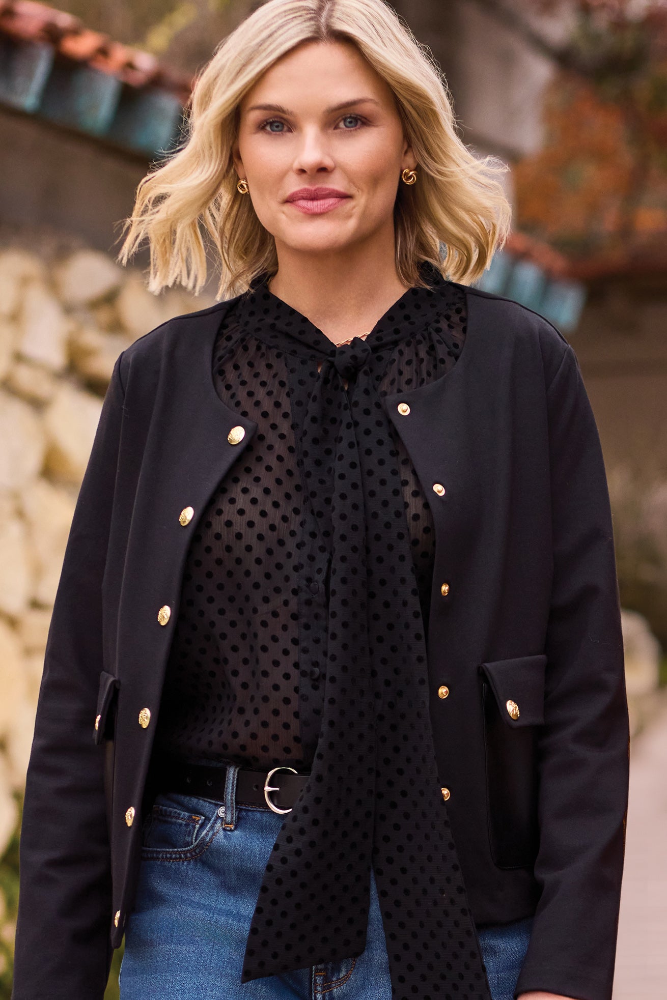 Black polka dot blouse worn under a black jacket with blue jeans, with a black belt, on person in an outdoor setting.