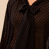 A close-up of a woman wearing a black sheer long-sleeve blouse with a black polka dot pattern and a large bow at the V-neck.
