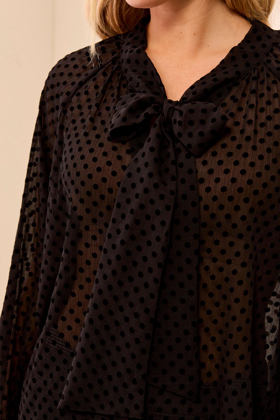 A close-up of a woman wearing a black sheer long-sleeve blouse with a black polka dot pattern and a large bow at the V-neck.