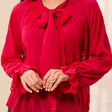 A woman wearing a bright pink long-sleeve blouse with a bow at the collar, ruffled cuffs, and a subtle polka dot pattern, paired with dark gray pants against a light beige background.