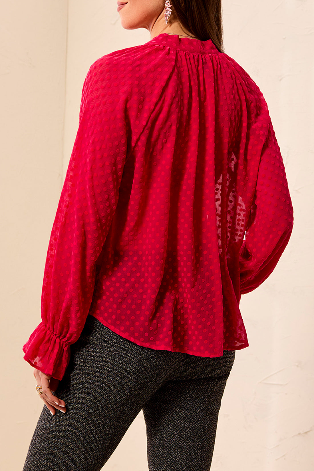 A woman wearing a bright pink long-sleeve blouse with a subtle polka dot pattern, shown from the back. The blouse features a gathered high neck and ruffled cuffs.