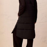 Person wearing a long black vest with a textured beige wall background