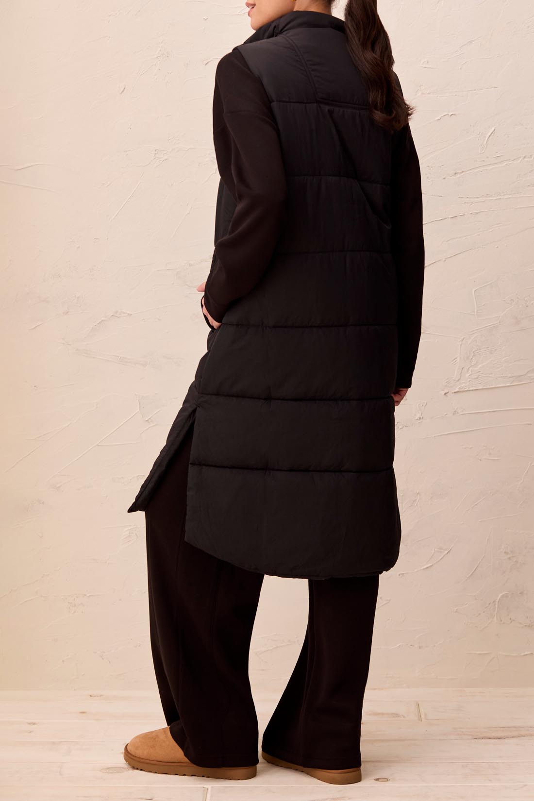 Person wearing a long black vest with a textured beige wall background