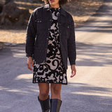 Woman walking outdoors wearing a black jacket, patterned dress, and black boots.