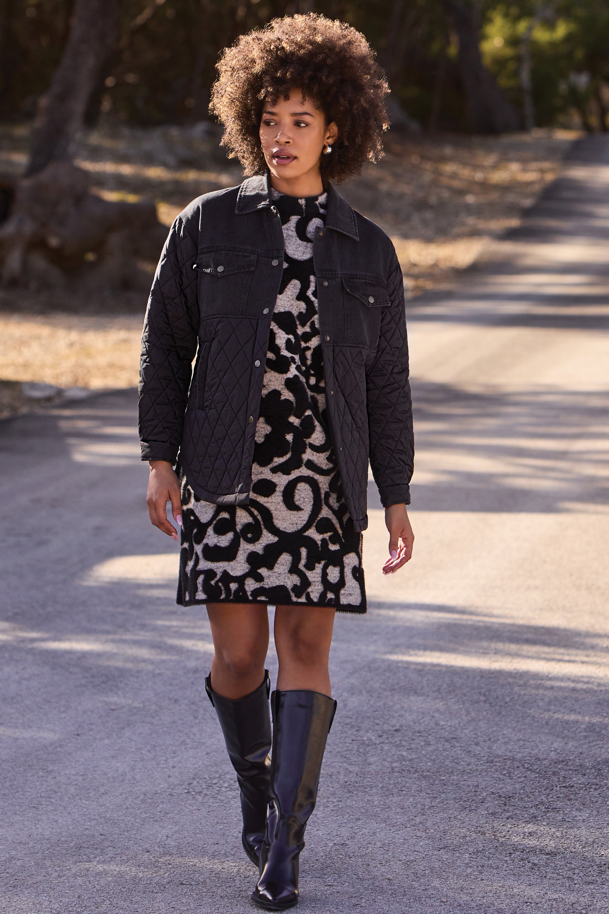 Woman walking outdoors wearing a black jacket, patterned dress, and black boots.