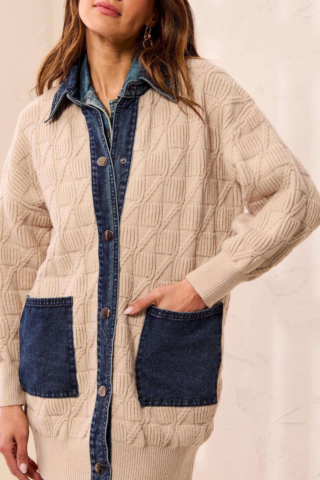Person wearing a beige cable knit sweater with blue denim pockets, placket and collar against a light background
