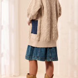 Person, shown from behind, wearing a beige cable knit sweater and blue skirt with brown boots against a white curtain background