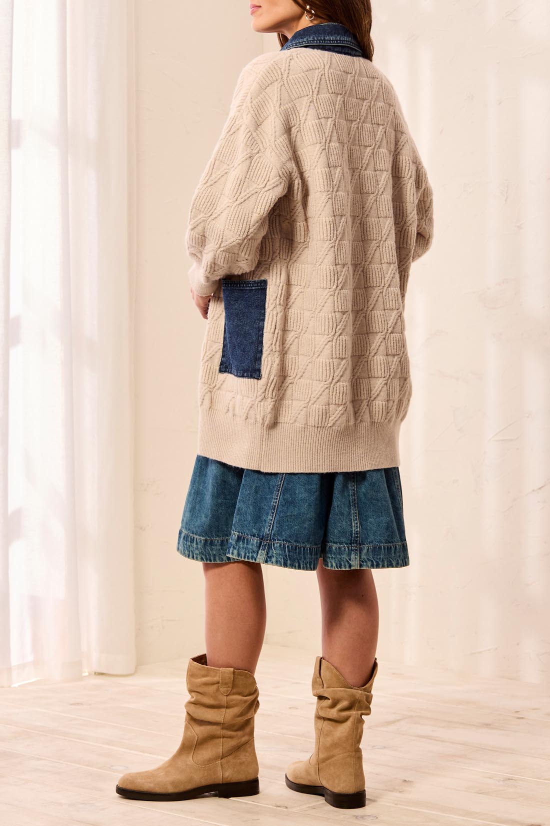 Person, shown from behind, wearing a beige cable knit sweater and blue skirt with brown boots against a white curtain background