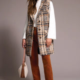 Woman wearing a patterned vest, white shirt, brown pants, and brown shoes against a plain background