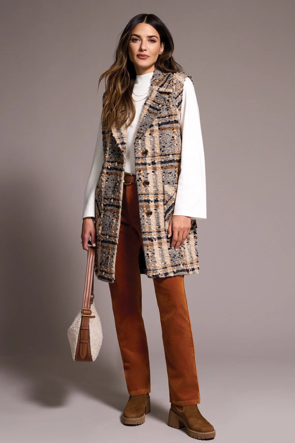 Woman wearing a patterned vest, white shirt, brown pants, and brown shoes against a plain background