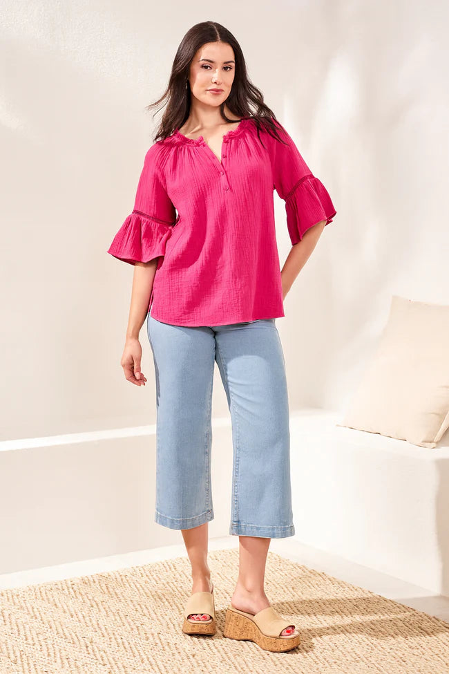 Woman wearing a pink blouse and light blue jeans standing in a room with a neutral background.