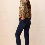 Woman wearing a patterned top and dark jeans against a beige background