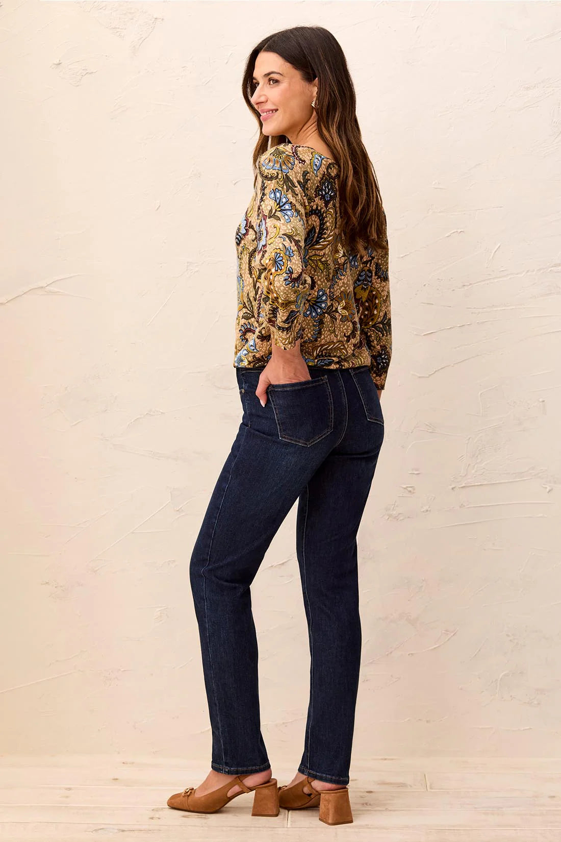 Woman wearing a patterned top and dark jeans against a beige background