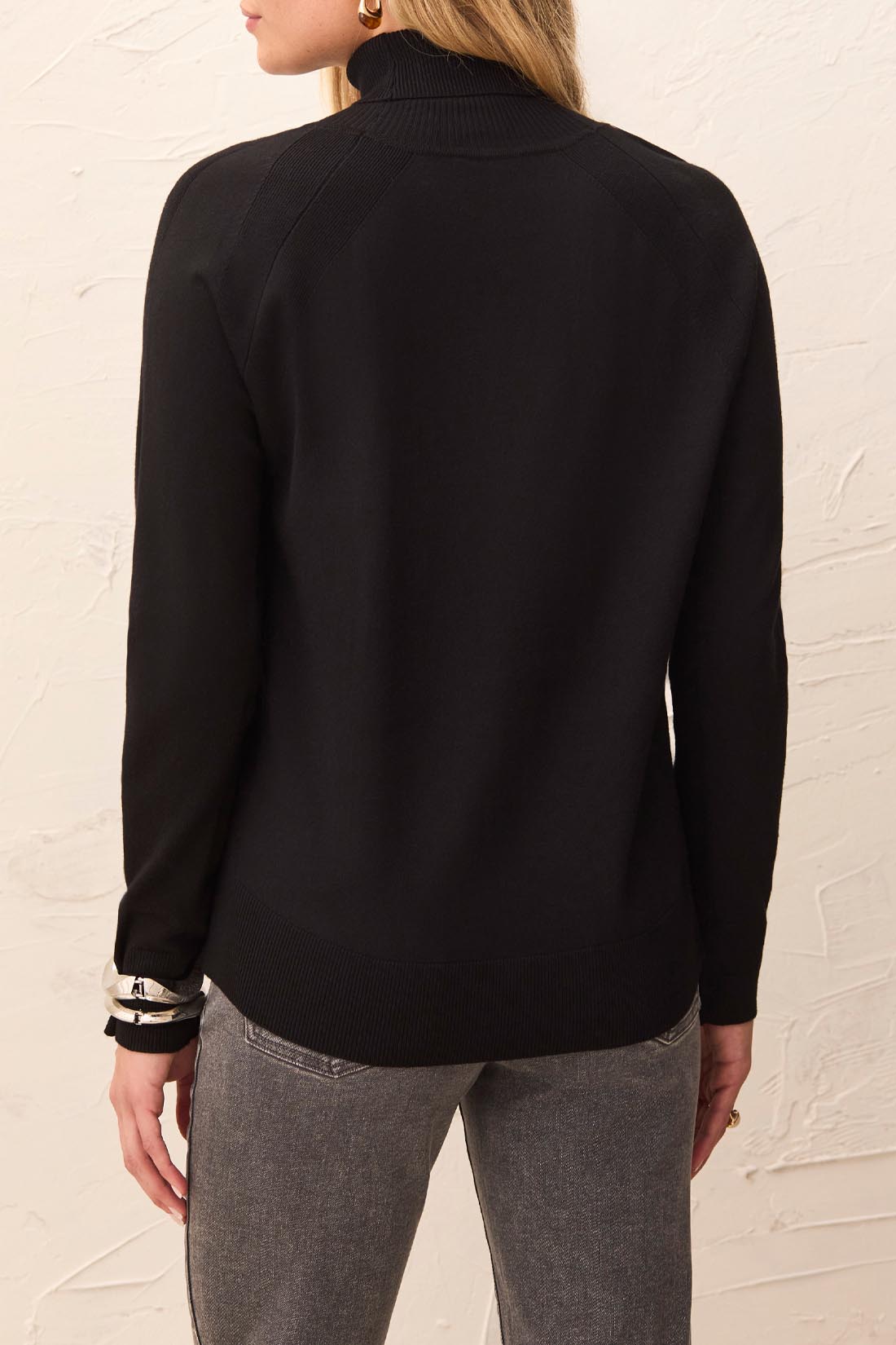 Person wearing a black long-sleeve top with a textured white wall background
