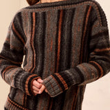 A woman wearing a vertical striped black, grey and orange sweater with grey jeans against a light backdrop.