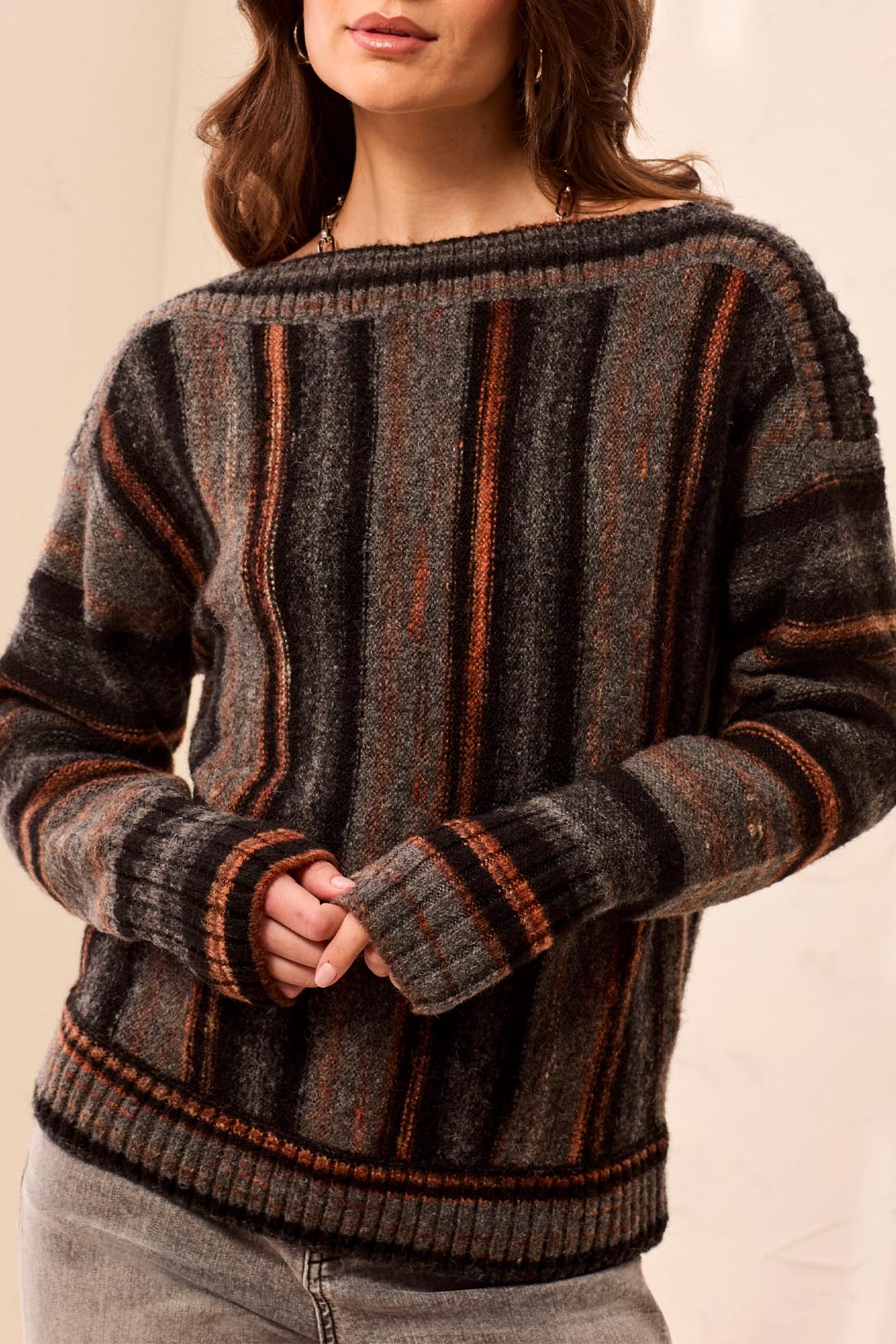 A woman wearing a vertical striped black, grey and orange sweater with grey jeans against a light backdrop.