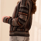 A woman, from a side profile, wearing a vertical striped black, grey and orange sweater with grey jeans against a light backdrop.