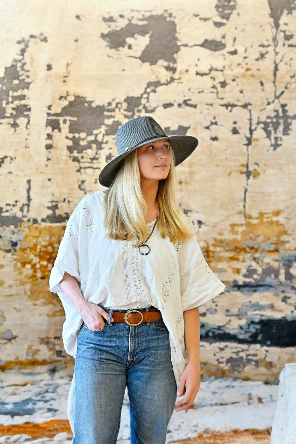Woman wearing a white blouse, blue jeans, and a gray hat against a textured wall.