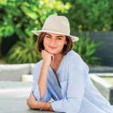 Woman wearing a light blue cardigan and beige hat sitting outdoors with greenery in the background