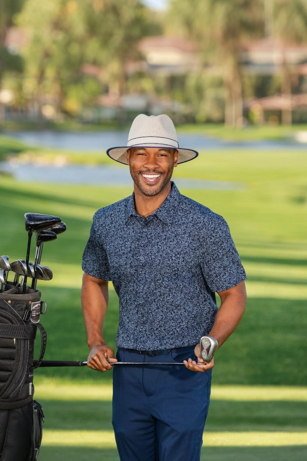 Man in a blue patterned shirt and white with navy trim hat holding a golf club on a golf course.