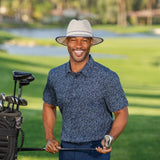 Man in a blue patterned shirt and white with navy trim hat holding a golf club on a golf course.