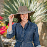 Woman wearing a denim shirt and wide-brimmed hat outdoors with plants in the background