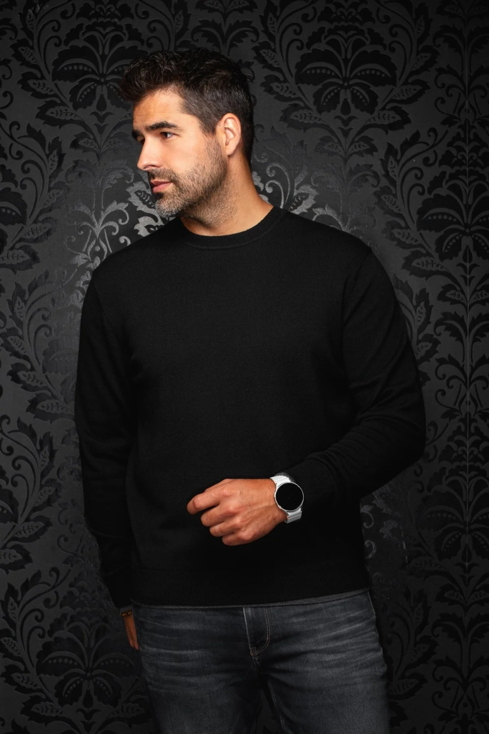Man wearing a black Au Noir LACAUNE cashmere sweater from Broderick's Clothing Co. against a dark patterned wall.