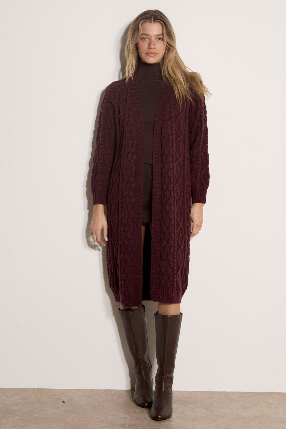Broderick's Clothing Co. burgundy cable knit midi cardigan worn by a model with brown boots against a white wall.