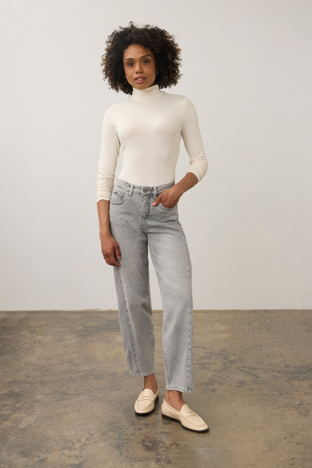 Model wearing a Broderick's Clothing Co. cream long sleeve mock neck top tucked into grey jeans with beige loafers.