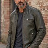 Broderick's Clothing Co. olive green leather bomber jacket with stand-up collar worn by a man against a brick wall.