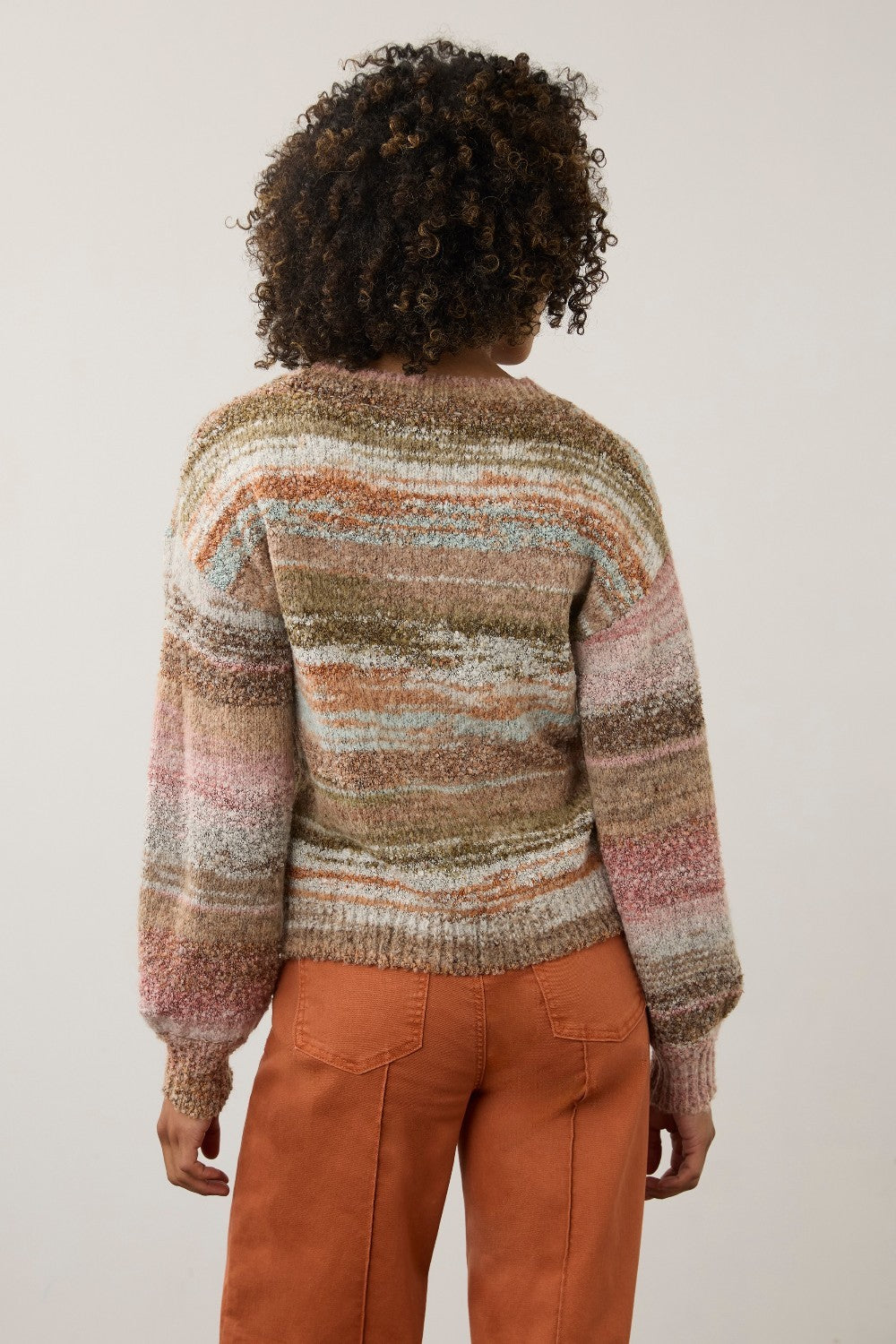 A person shown from the back wearing a fuzzy striped knit sweater with horizontal bands of beige, brown, pink, and muted blue, paired with rust-orange pants, against a plain light beige studio background.