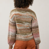 A person shown from the back wearing a fuzzy striped knit sweater with horizontal bands of beige, brown, pink, and muted blue, paired with rust-orange pants, against a plain light beige studio background.