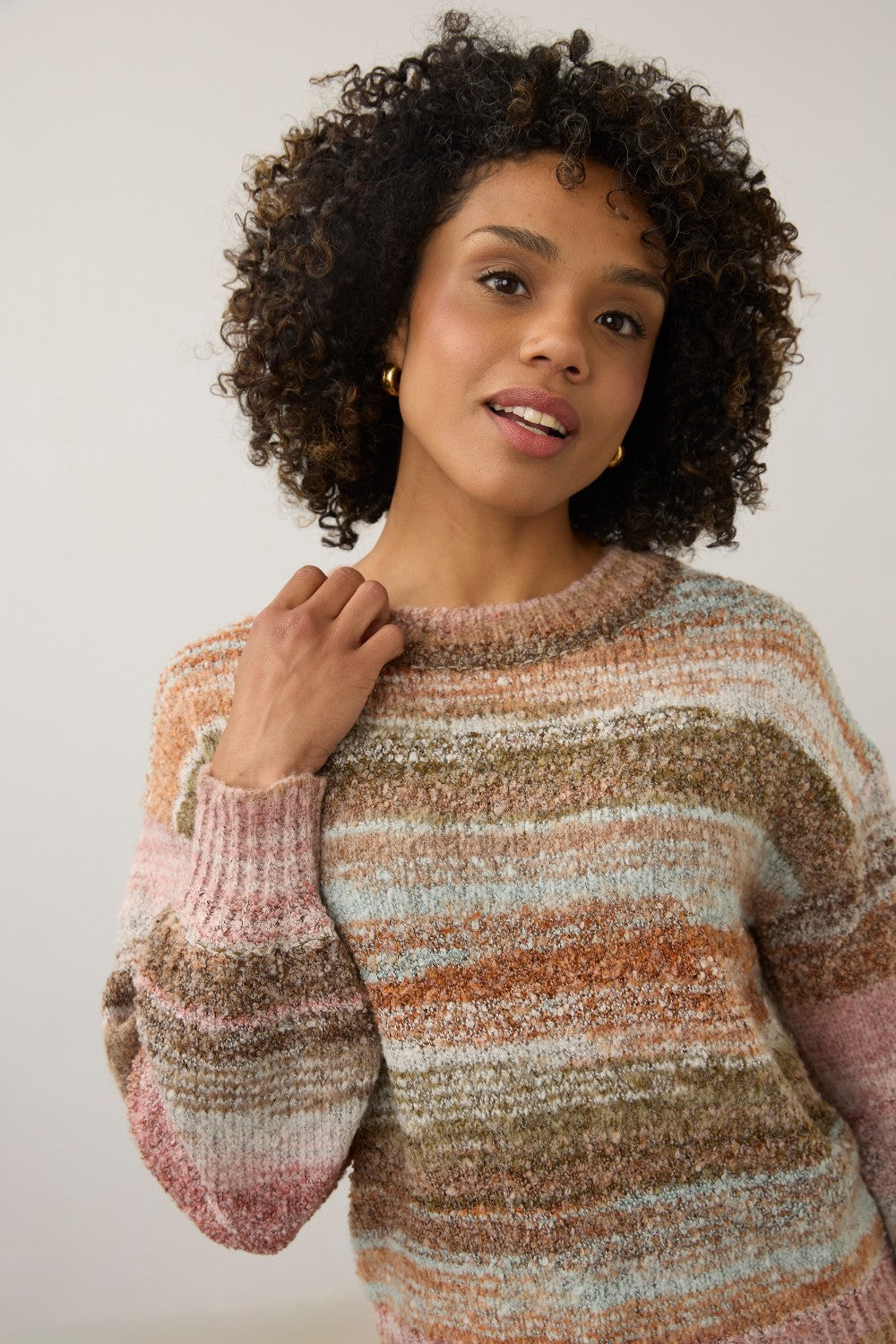 A woman with curly dark hair wearing a chunky, marled knit sweater with horizontal stripes in dusty pink, brown, beige, and light blue, posed against a plain light gray background.