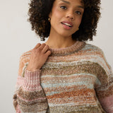 A woman with curly dark hair wearing a chunky, marled knit sweater with horizontal stripes in dusty pink, brown, beige, and light blue, posed against a plain light gray background.