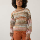 A woman with curly dark hair wearing a multicolor striped knit sweater and rust-orange pants, standing against a plain light gray studio background.