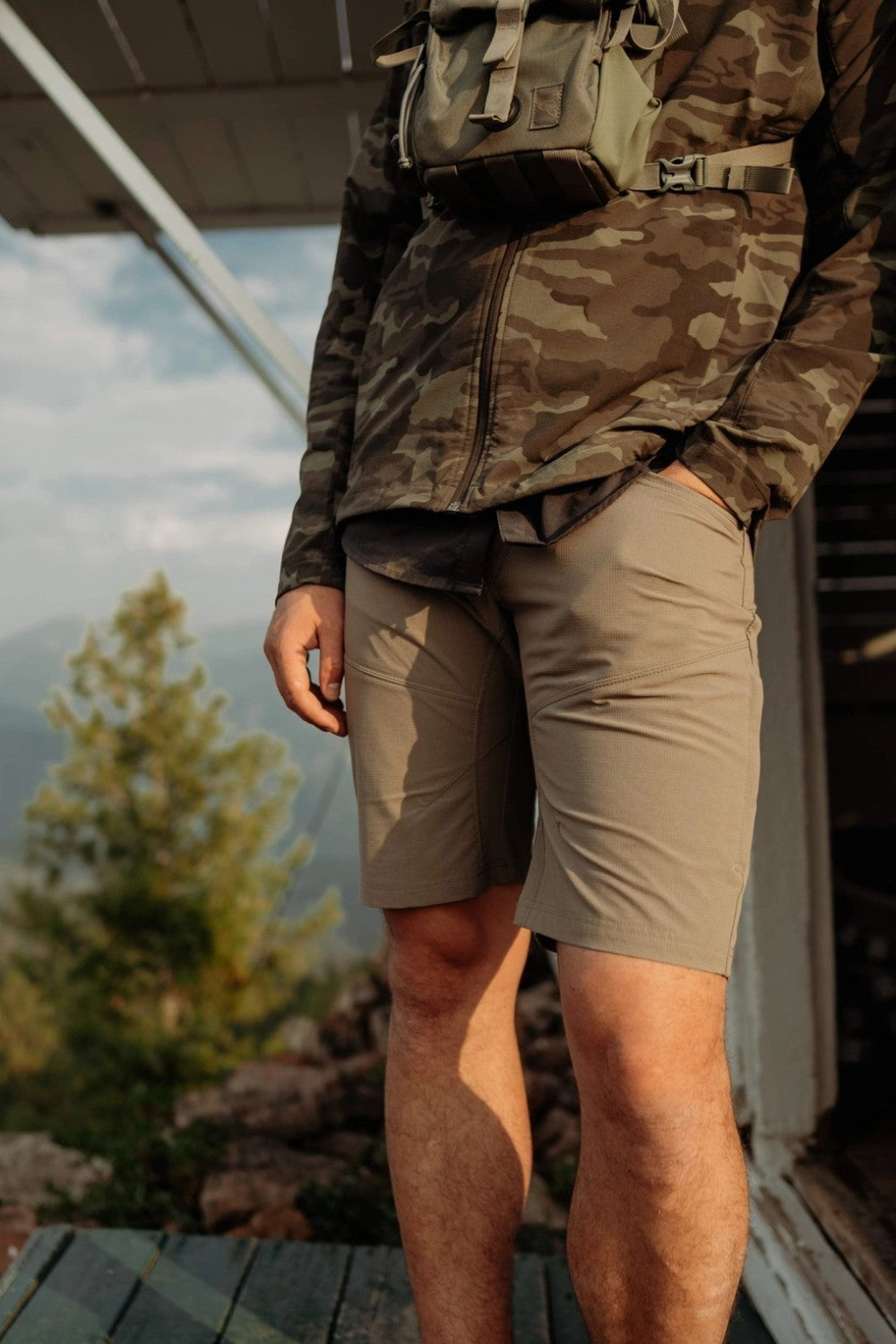 Person wearing camouflage shirt and beige shorts standing outdoors with a scenic background.