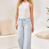 Woman wearing a white sleeveless top and light blue jeans standing in a room with beige cushions and a plant.