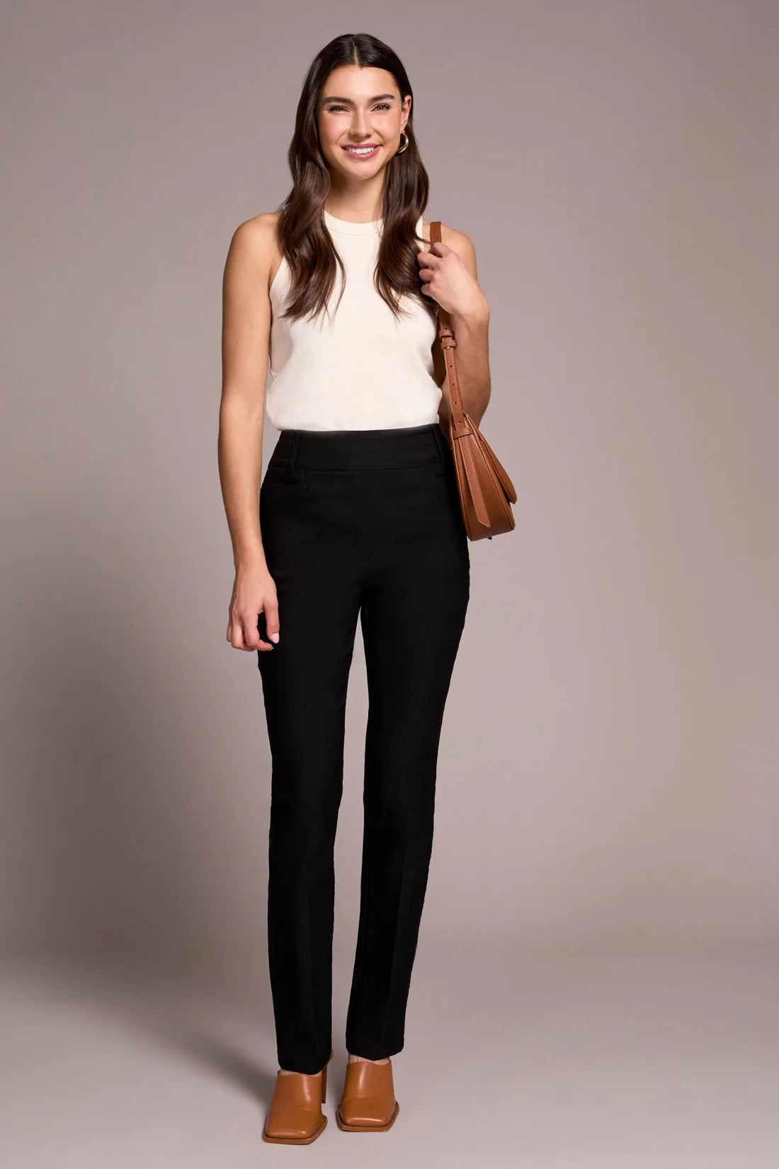 Woman wearing a white sleeveless top and black pants, holding a brown handbag against a plain background