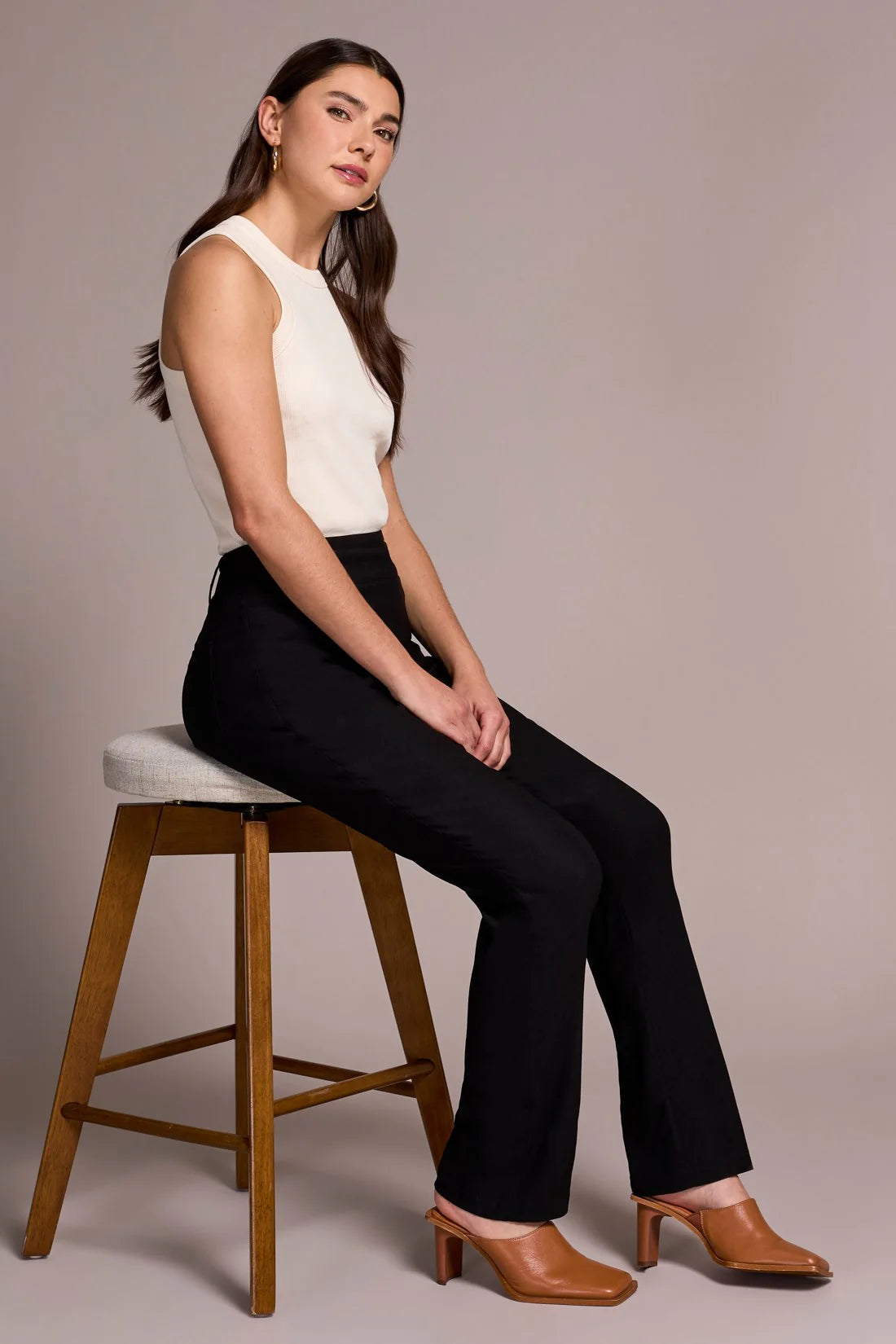 Woman sitting on a wooden stool wearing a white top and black pants against a plain background