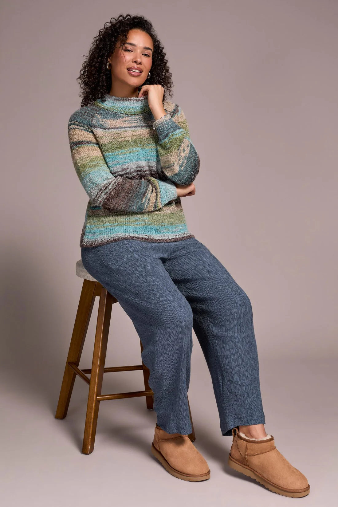 Woman wearing a colorful sweater and blue jeans sitting on a wooden stool against a plain background