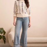Woman wearing a cream sweater with black leaf patterns, light blue jeans, and brown boots in a room with a beige sofa and plant.