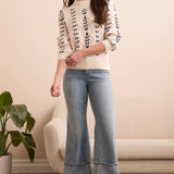 Woman wearing a patterned sweater, jeans, and boots in a room with a plant and sofa.